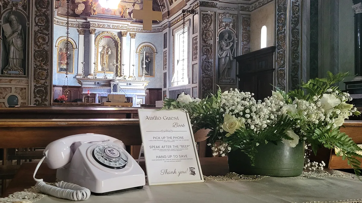 Téléphone vintage blanc et cadre livre d’or audio installés dans une église, au milieu des fleurs