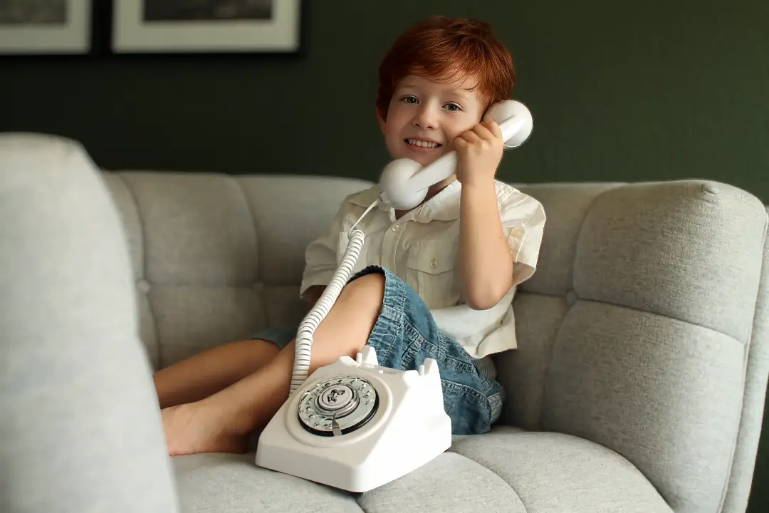 Enfant souriant utilisant un téléphone rétro blanc, assis sur un canapé.