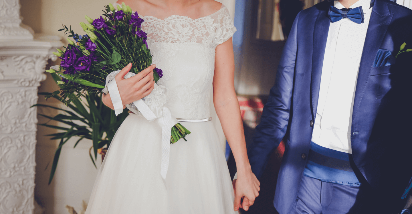 Mariée en robe blanche et marié en costume bleu, main dans la main lors de leur mariage, bouquet de fleurs violettes en main.